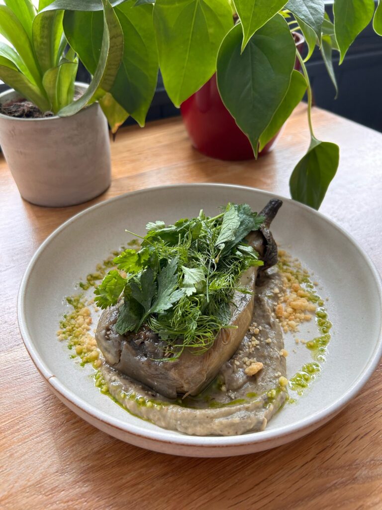 Aubergine farcie gourmande sur purée, garnie d'herbes fraîches (menthe, aneth, coriandre), huile verte et crumble. Table en bois, plantes.