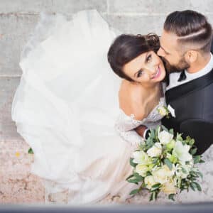 Mariés souriants, le marié embrasse la joue de la mariée en robe blanche. Elle tient un bouquet de fleurs blanches et vertes.