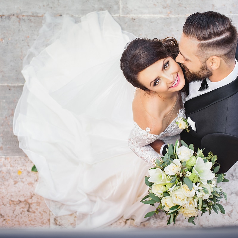 Mariés souriants, le marié embrasse la joue de la mariée en robe blanche. Elle tient un bouquet de fleurs blanches et vertes.