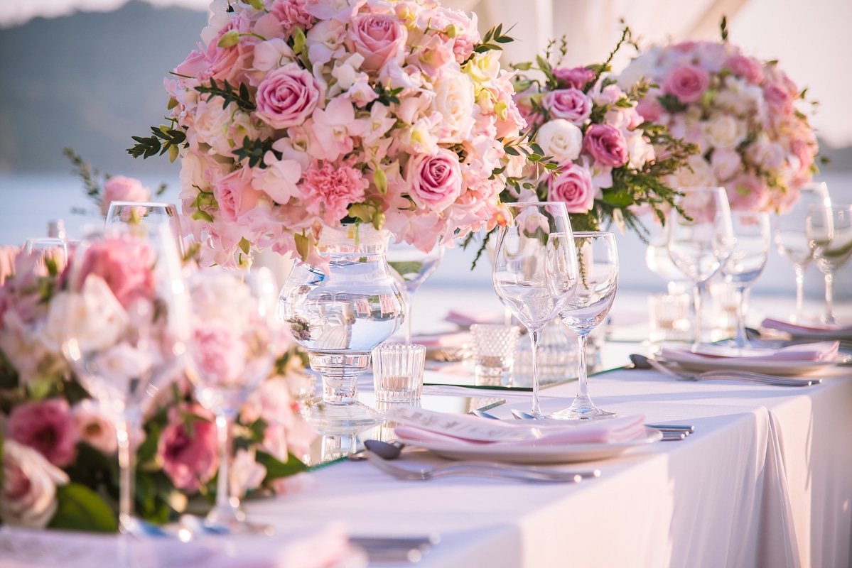 Table de mariage élégante avec centres de table floraux roses, verres et assiettes. Décor extérieur raffiné, ambiance douce.