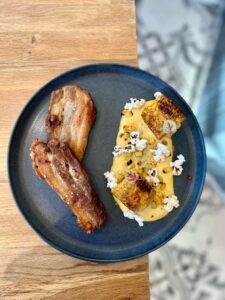Deux morceaux de poitrine de porc rôtie salée, avec purée de maïs, maïs grillé, pop-corn et micro-pousses sur une assiette.