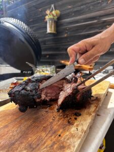 Un homme tranche une viande fumée juteuse sur une planche à découper en bois, avec un grand barbecue et de la fumée en arrière-plan.