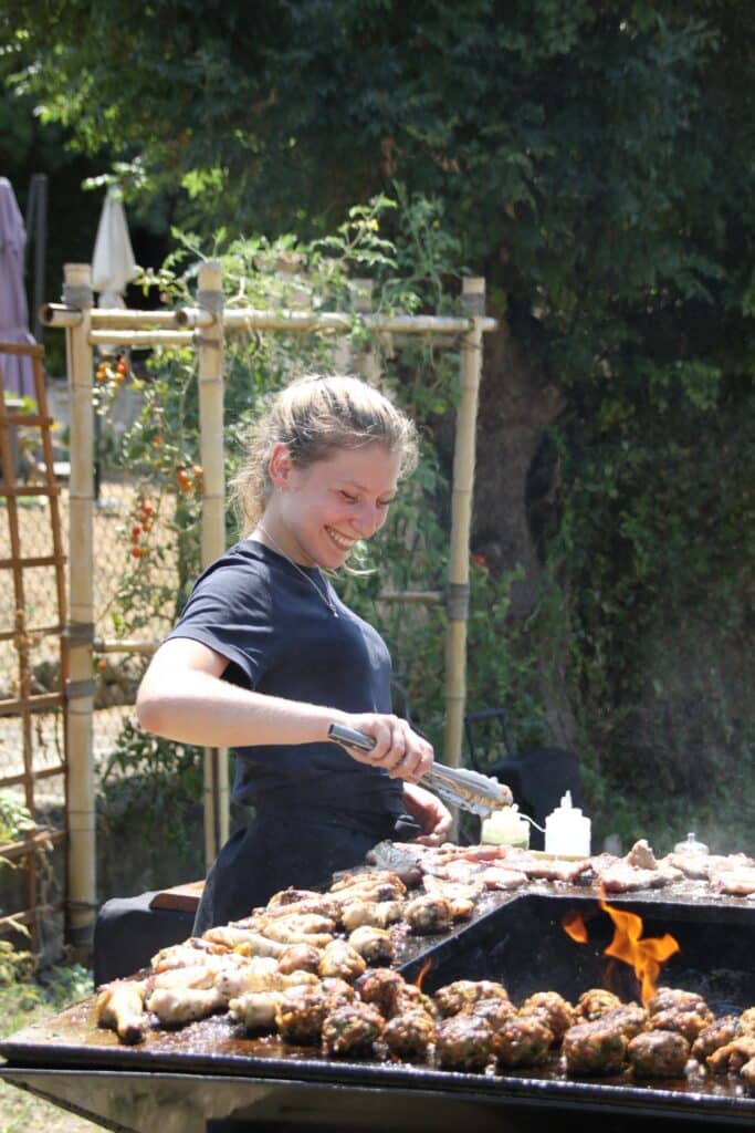 Femme souriante grillant viande et boulettes avec des pinces sur un grand barbecue extérieur, flammes vives.