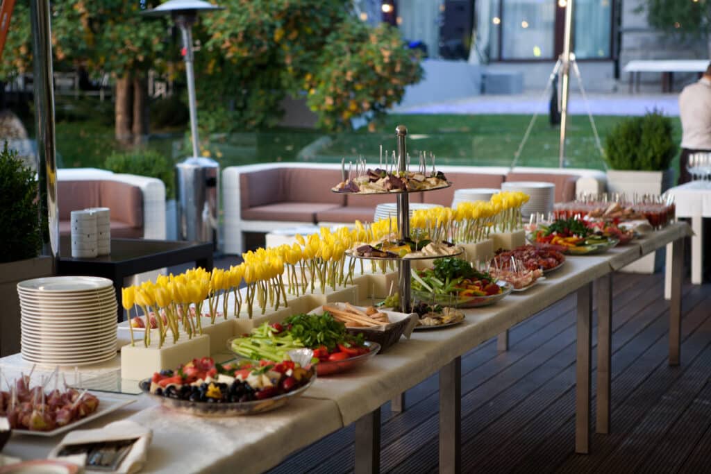 Grand buffet extérieur élégant garni d'apéritifs: fromages, charcuteries, crudités, fruits variés. Tulipes jaunes et assiettes empilées.