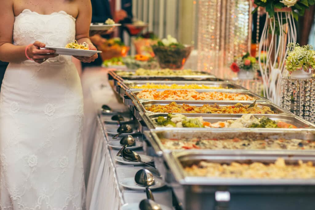 Mariée en robe blanche prenant un plat à un buffet traiteur. Plusieurs chafing dishes présentent des plats chauds variés, décorations de mariage.