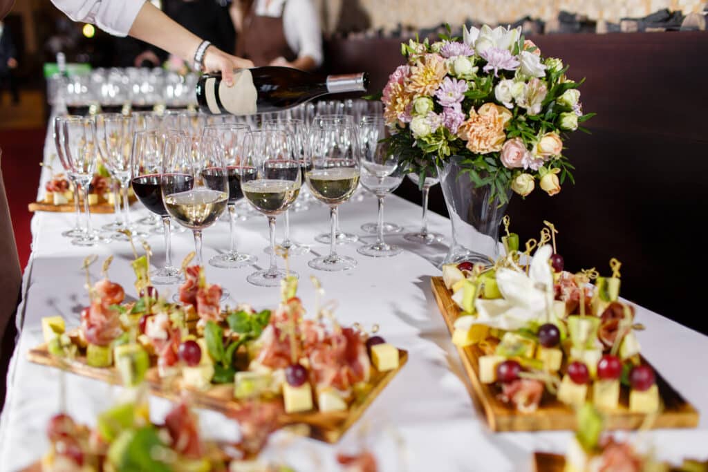 Main versant du vin rouge et blanc dans des verres sur une table de réception. Canapés, fromages et fruits, ainsi qu'un bouquet floral coloré.
