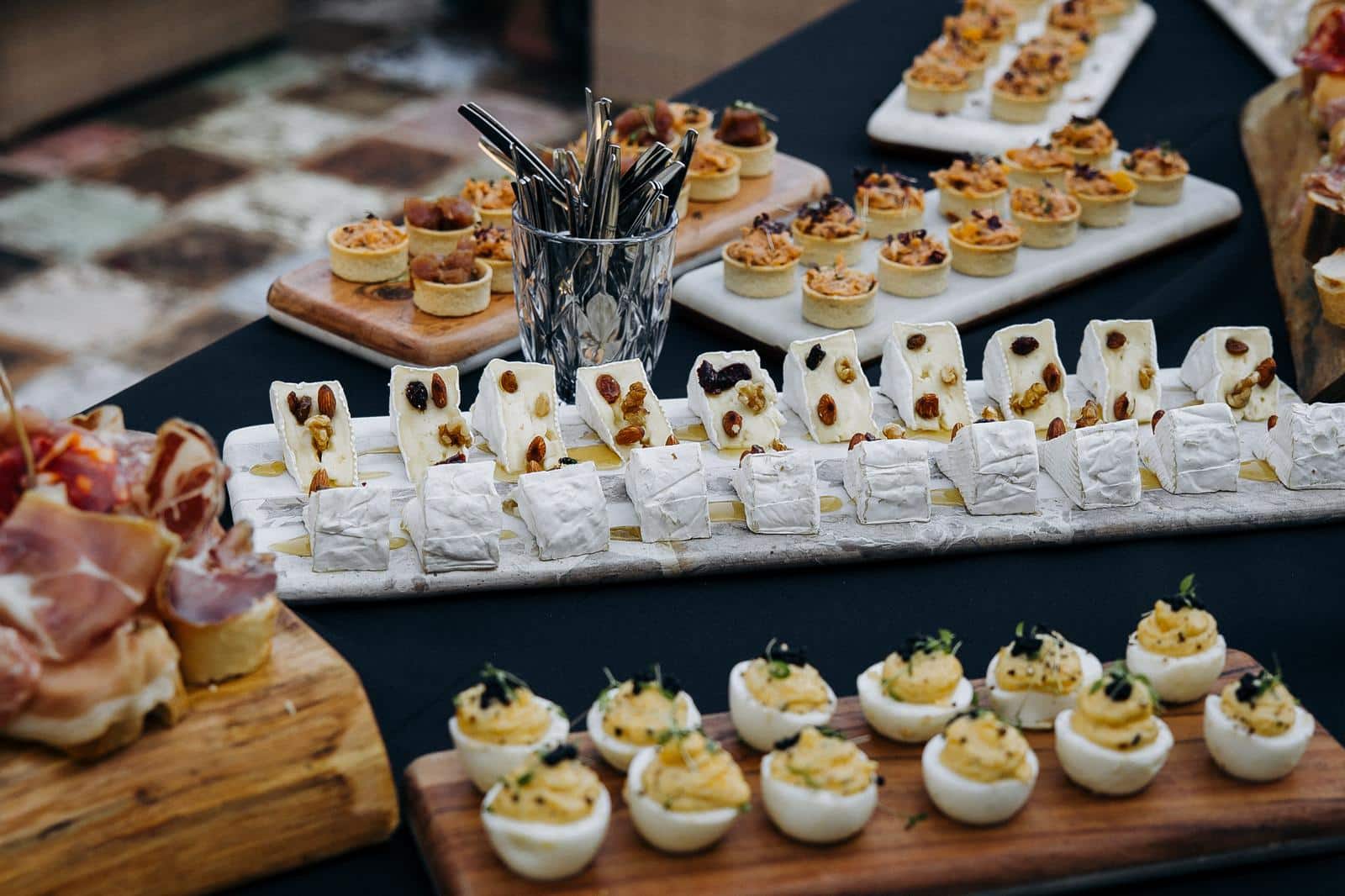 Table de traiteur variée: fromages brie aux noix et miel, charcuterie, œufs mimosa au caviar, et mini-tartes salées. Buffet apéritif élégant.