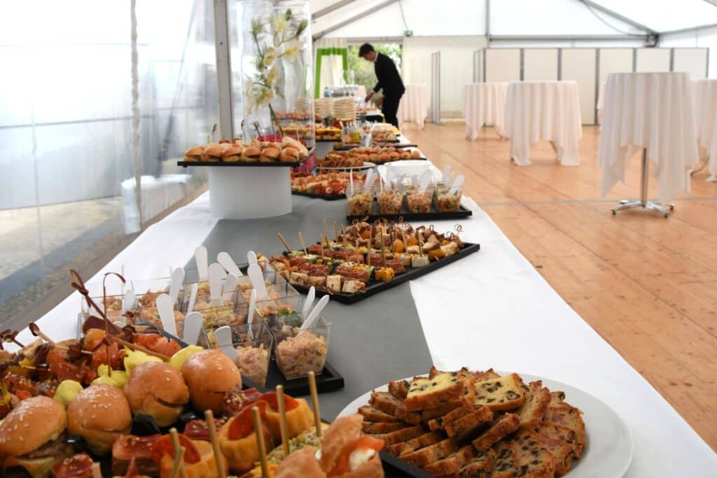 Buffet de traiteur avec amuse-bouches variés (mini-burgers, brochettes, verrines) et cakes dans une tente événementielle. Un serveur prépare les tables.