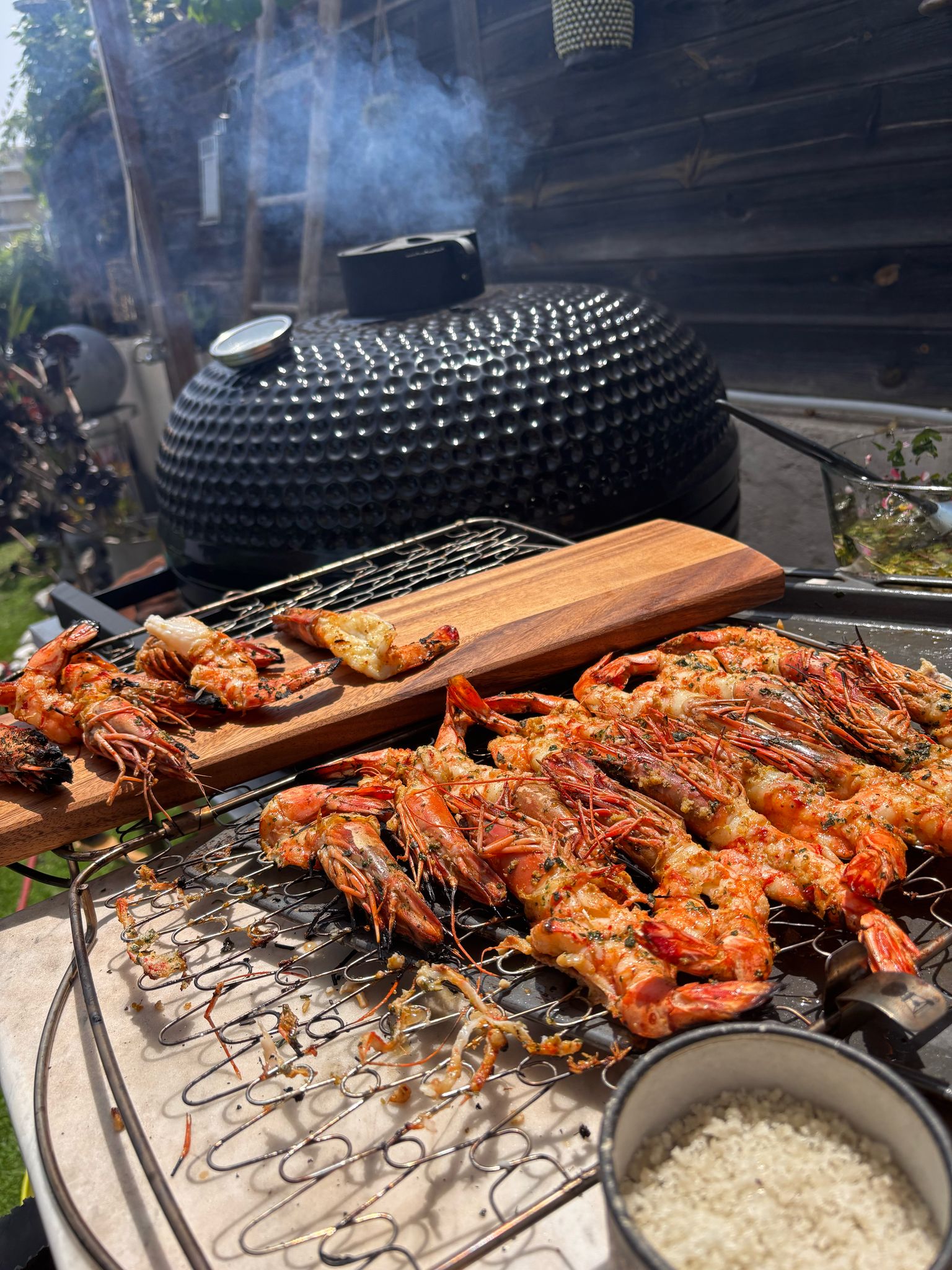 Grosses crevettes grillées assaisonnées sur un barbecue kamado noir fumant en plein air, avec planche en bois et sel.