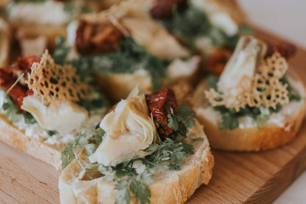Crostini élégants sur planche en bois, garnis d'artichauts, tomates séchées, herbes fraîches et tuiles de fromage croustillantes.