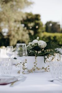 Décor de table élégant en extérieur avec centre de table de roses blanches, feuillage vert et guirlande dorée, verres raffinés.