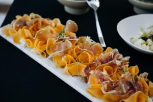 Longue assiette de fines tranches de melon, jambon de Parme, fromage frais et herbes, sur une table noire.