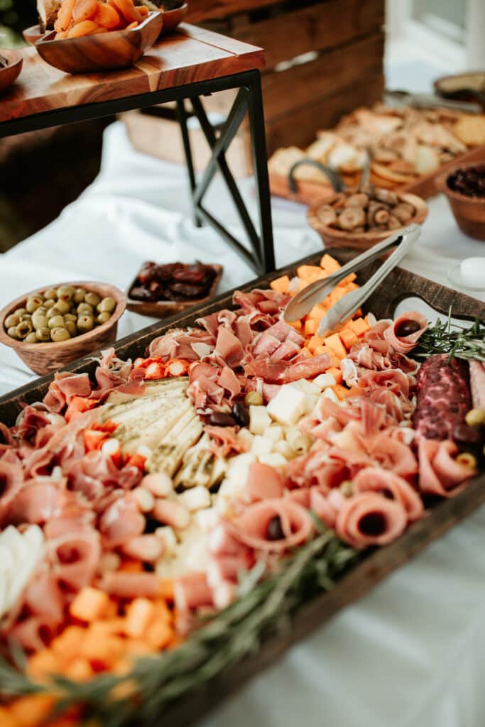 Grande planche de charcuterie garnie de jambon, salami, fromages cubés, olives et romarin. Bols d'abricots secs et snacks en arrière-plan.