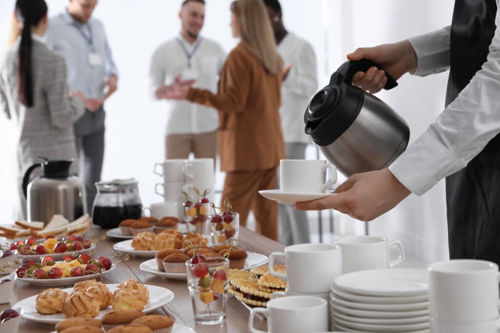 Main versant du café dans une tasse sur un buffet de réunion. Table garnie de pâtisseries, fruits, sandwichs. Participants en fond.