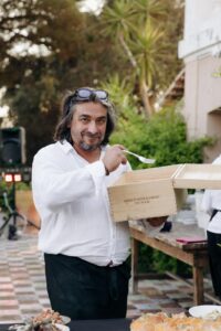 Homme souriant en chemise blanche et tablier, tenant une boîte de vin « NERO D'AVOLA SYRAH DOC SICILIA » et une fourchette lors d'un événement.