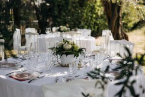 Table de réception élégante en extérieur avec centre de table de roses blanches, verrerie en cristal, et chaises blanches, dans un cadre verdoyant.