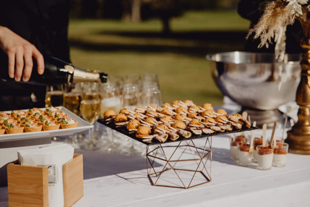 Table de traiteur extérieur: personne servant du champagne, assortiment de canapés, mini-burgers, desserts. Napkins 'de la Gastronomie'.