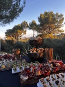Grand buffet extérieur au coucher du soleil avec charcuterie, fromages aux noix, pains et amuse-bouches, devant des pins illuminés.
