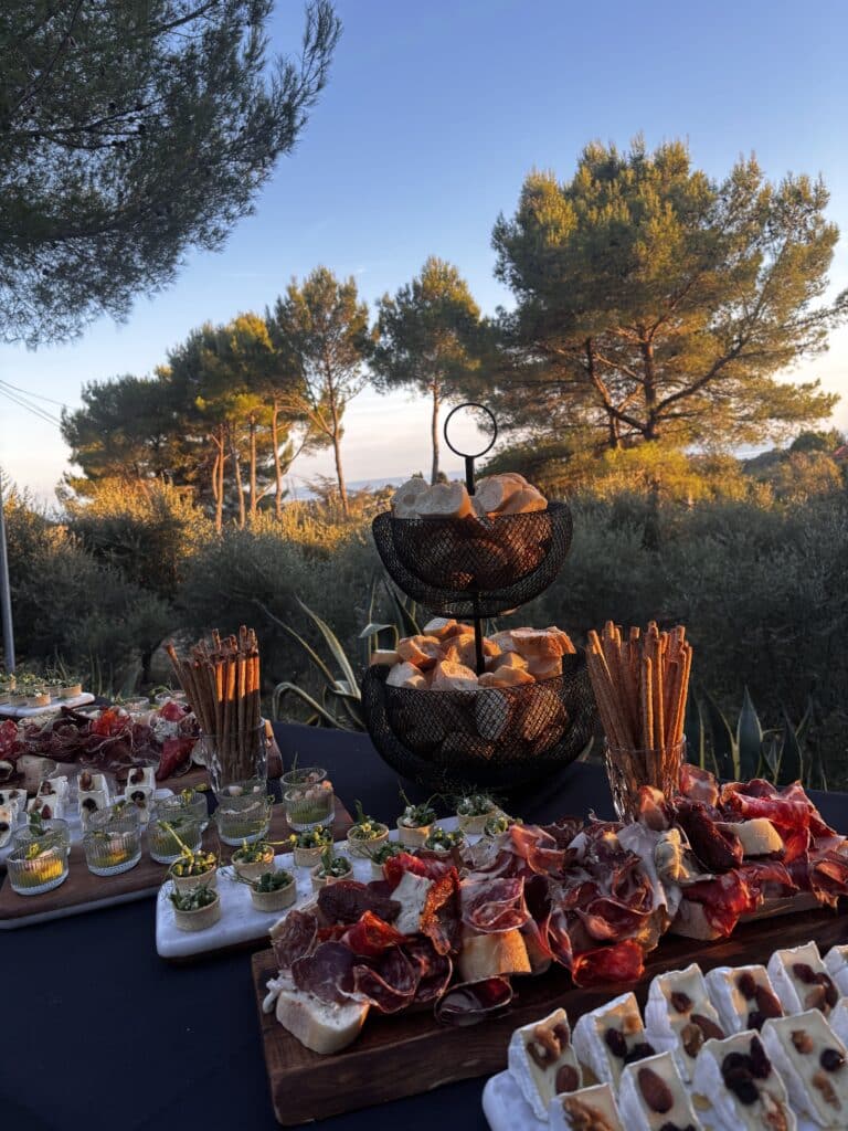 Grand buffet extérieur au coucher du soleil avec charcuterie, fromages aux noix, pains et amuse-bouches, devant des pins illuminés.