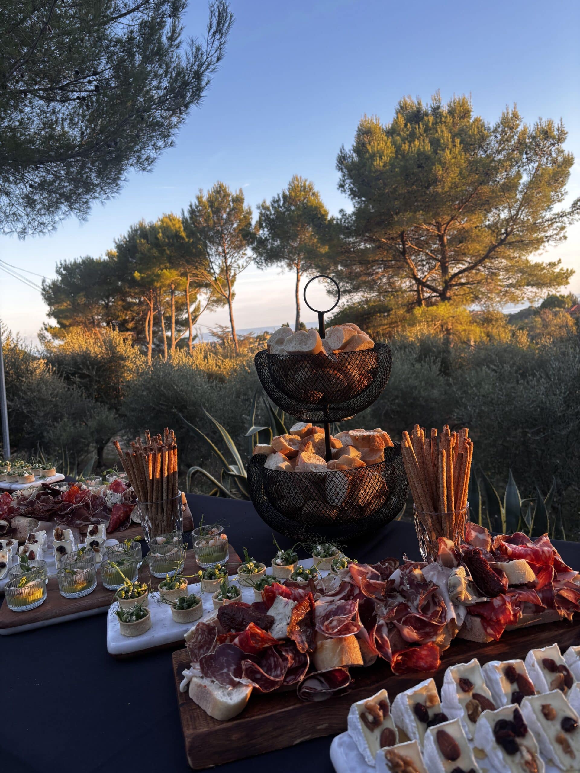 Grand buffet extérieur au coucher du soleil avec charcuterie, fromages aux noix, pains et amuse-bouches, devant des pins illuminés.