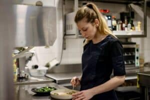Jeune cheffe blonde concentrée, en veste de cuisine bleue, dressant un plat avec précision dans une cuisine professionnelle moderne.