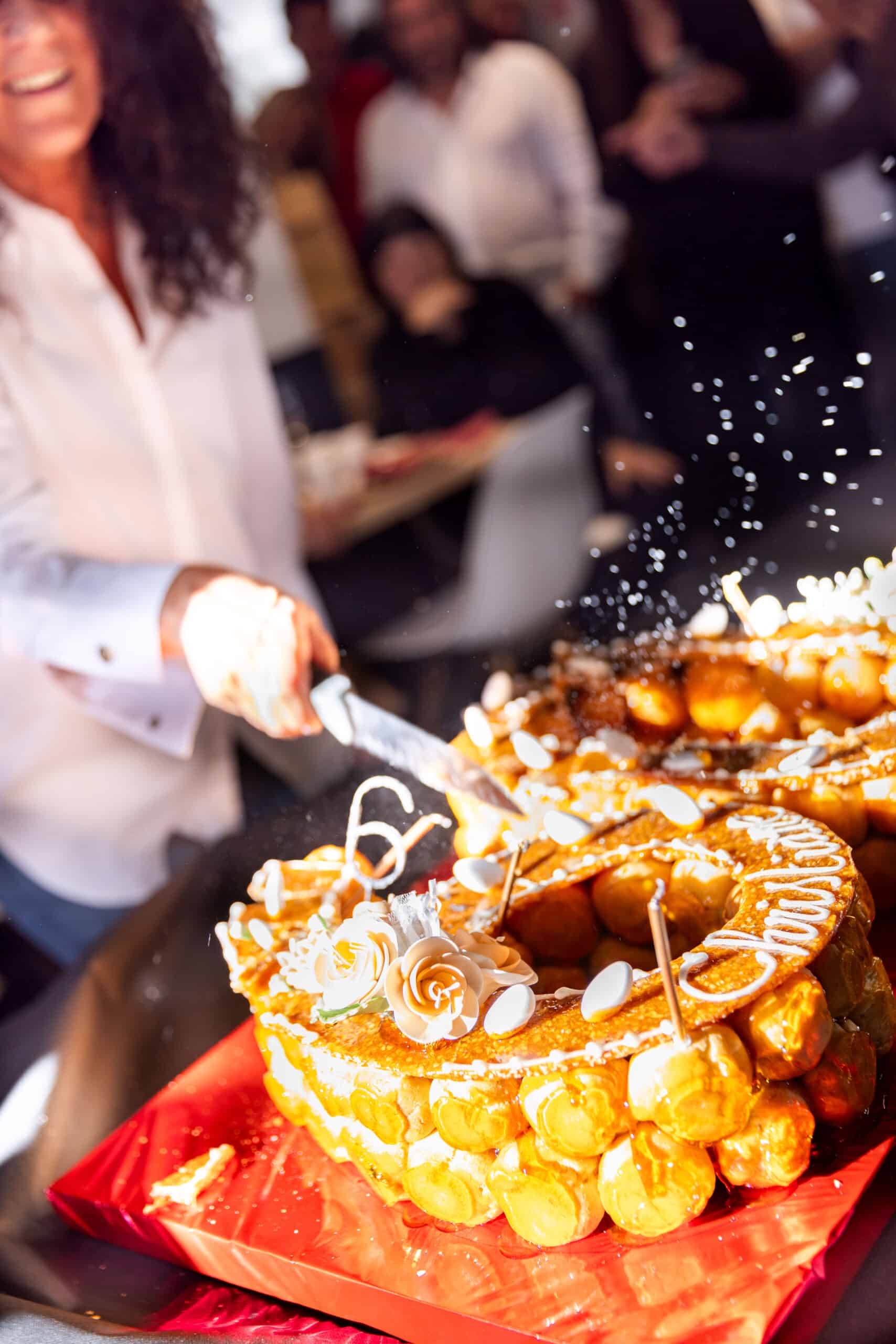 Femme souriante coupant un croquembouche doré, décoré de fleurs et du chiffre "6". Des éclats de sucre volent, ambiance joyeuse de fête.