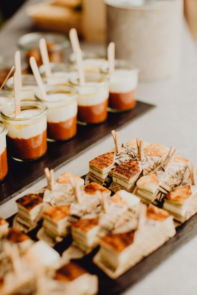 Buffet traiteur: verrines bicolores avec cuillères en bois et mini-sandwichs empilés, emballés dans du papier à motifs "LATEST NEWEST PATTERN".