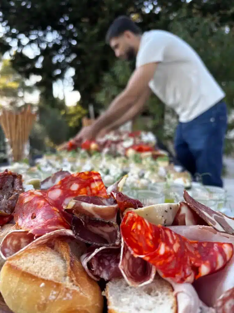 Ambiance apéritif extérieur avec gros plan sur charcuteries, pains et fromages, un homme arrangeant le buffet en arrière-plan.
