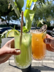 Deux mains trinquent des cocktails d'été frais : vert (citron vert, menthe, paille biodégradable) et orange (agrume séché). Terrasse ensoleillée.