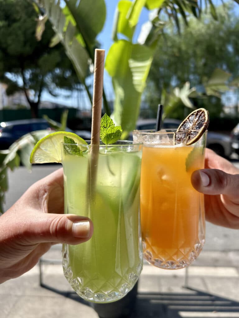 Deux mains trinquent des cocktails d'été frais : vert (citron vert, menthe, paille biodégradable) et orange (agrume séché). Terrasse ensoleillée.