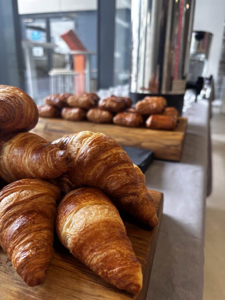 Croissants dorés empilés sur une planche en bois lors d'un buffet petit-déjeuner avec viennoiseries en arrière-plan.