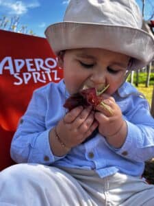 Bébé mangeant du jambon et de la roquette devant un panneau Aperol Spritz.