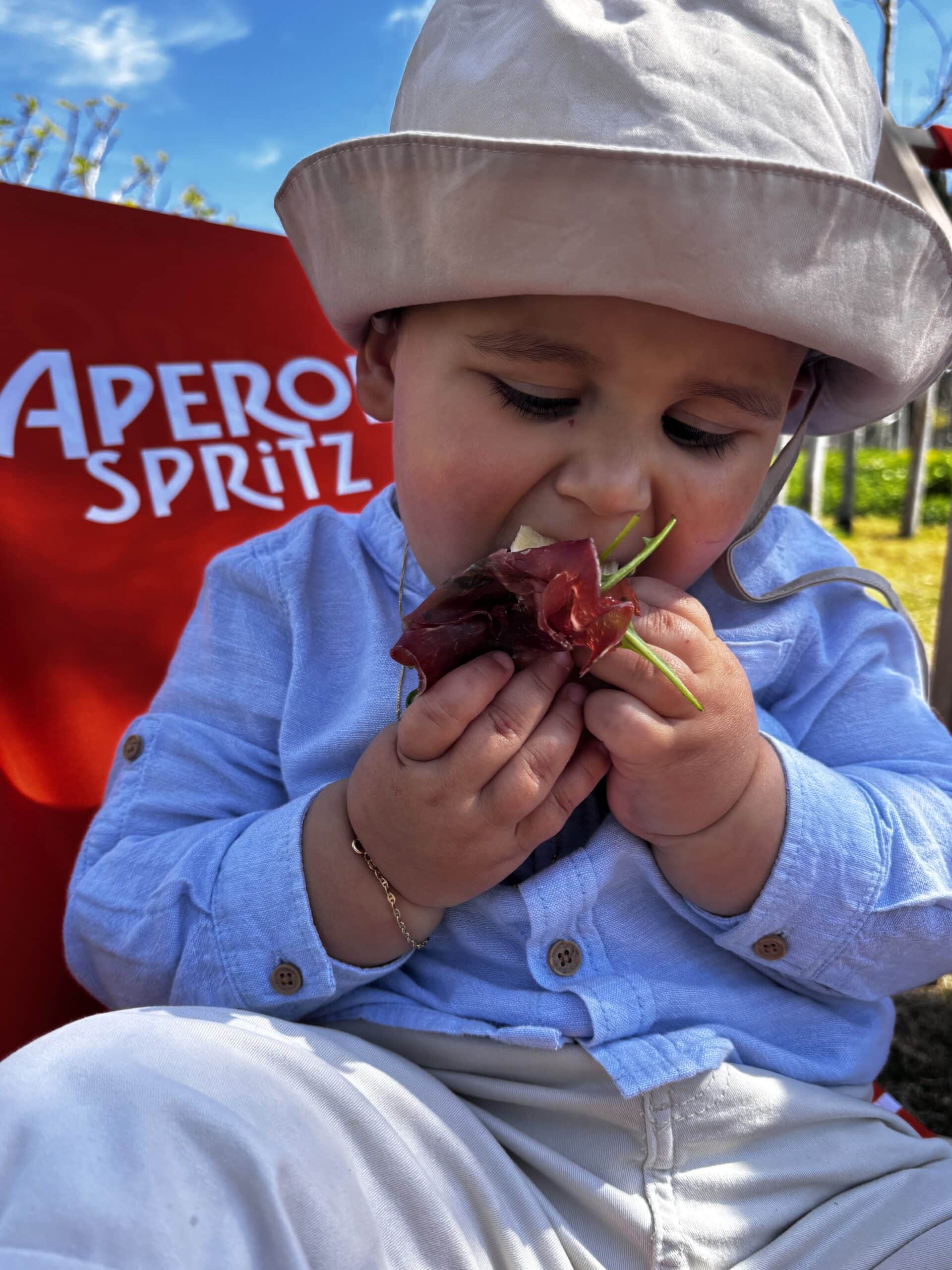 Bébé mangeant du jambon et de la roquette devant un panneau Aperol Spritz.