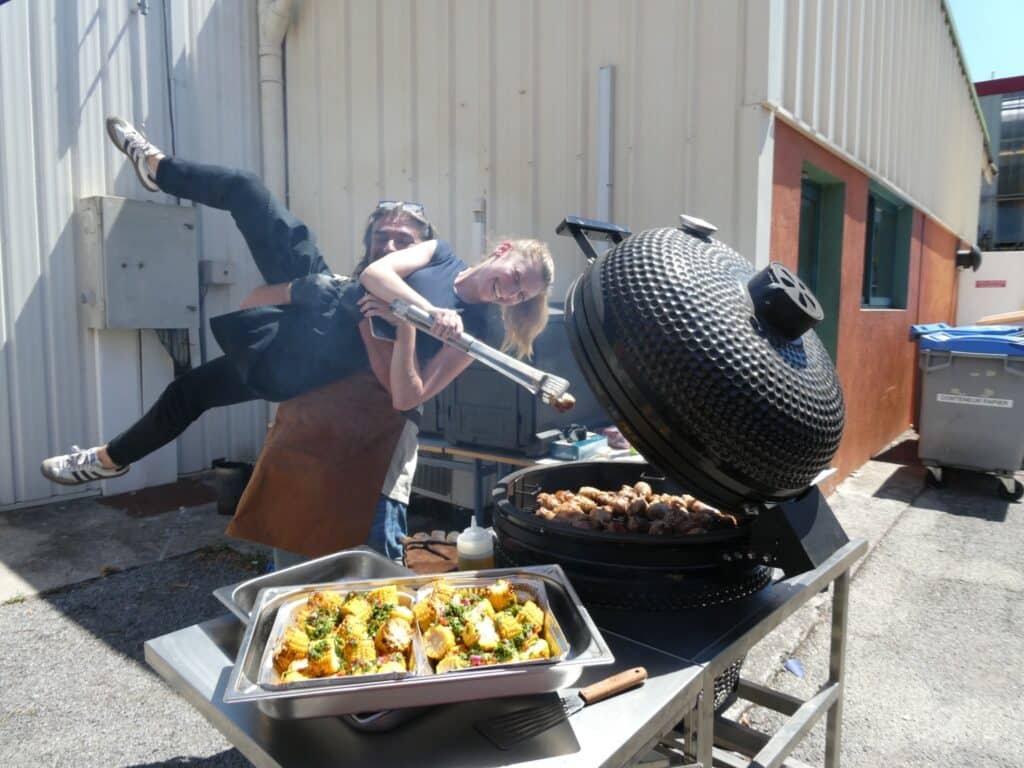 Barbecue Kamado : Cuisson Festive et Conviviale en Duo Duo joyeux cuisinant au barbecue Kamado noir, maïs grillé en plateau et ambiance festive en extérieur.
