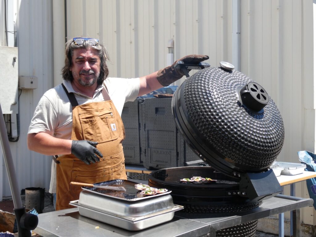 Barbecue Kamado : Cuisson de Saint-Jacques par un Chef Chef barbu en tablier brun grillant des coquilles Saint-Jacques sur un barbecue kamado noir ouvert.
