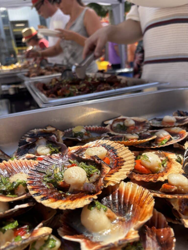 Coquilles Saint-Jacques grillées aux herbes sur un étal de marché extérieur avec foule.