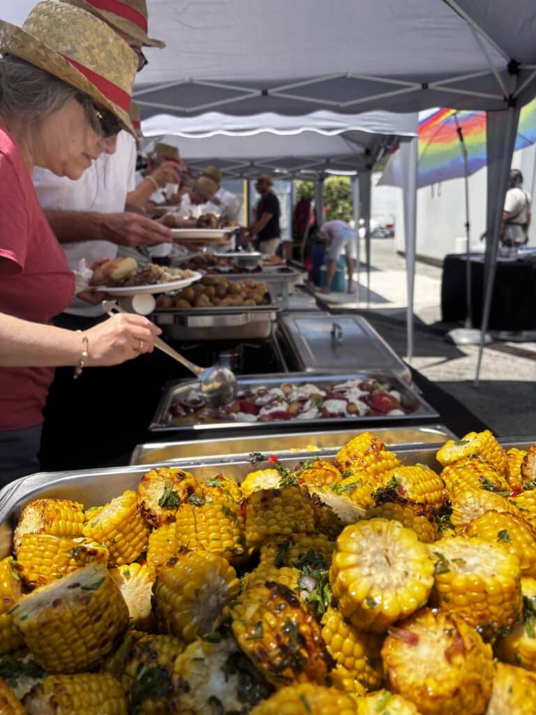 Maïs Grillé et Buffet de Fête en Extérieur sous Tente Gros plan de maïs grillé aux herbes lors d'un buffet festif en extérieur avec des personnes se servant.