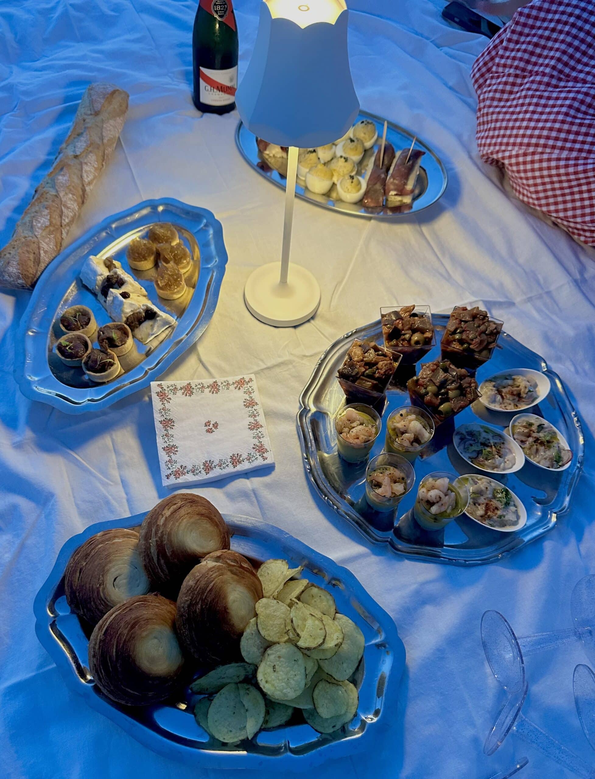 Pique-nique chic : champagne G.H. Mumm, baguette, croissants et verrines apéritives sur une nappe blanche éclairée.