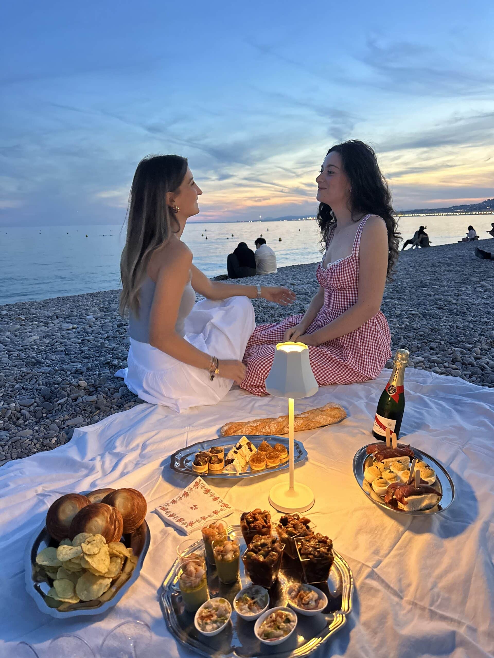 Deux amies savourent un pique-nique chic au coucher du soleil sur une plage de galets avec tapas et champagne.